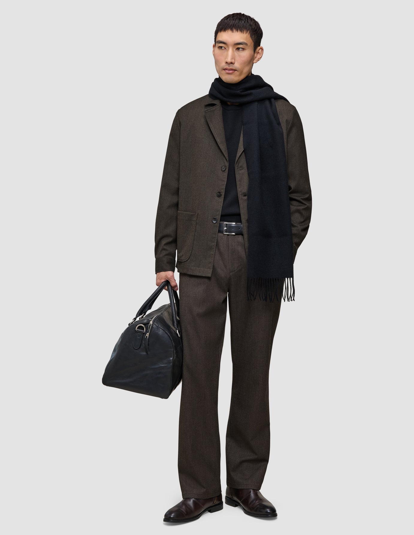 En mann i mørkebrune Atelier Pants Relaxed, svart genser og skjerf, som holder en svart skinnreiseveske.